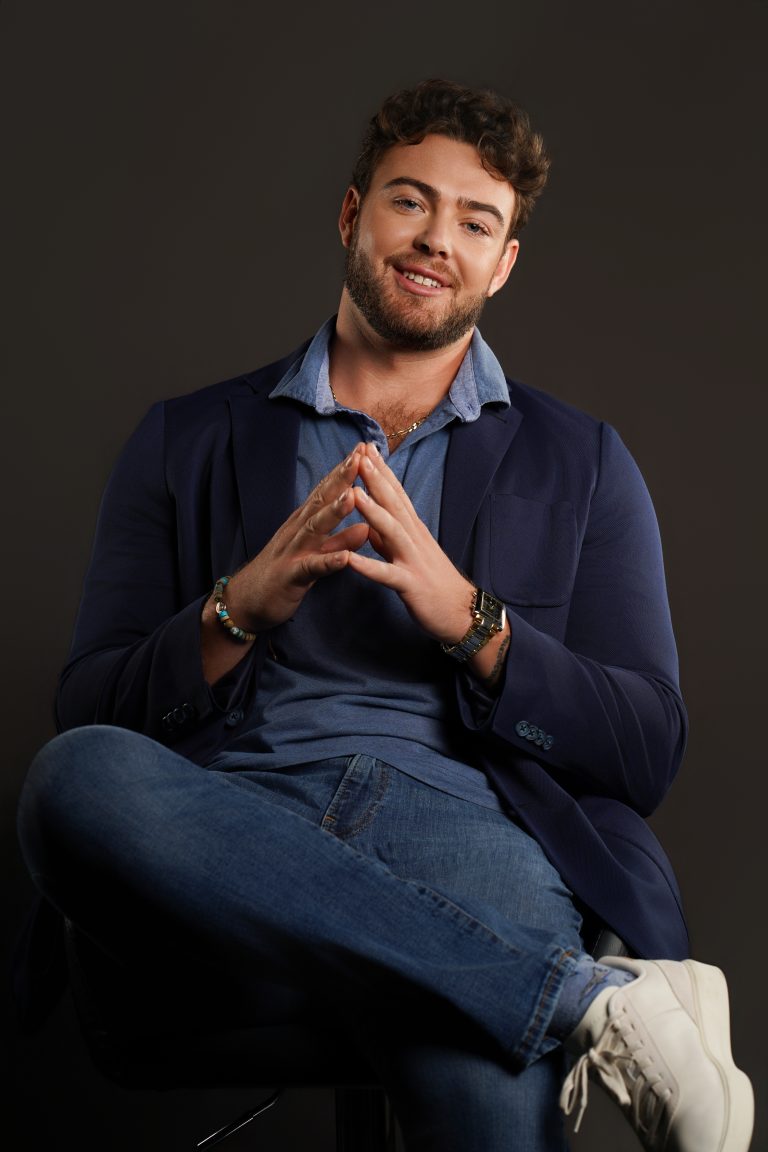 Jake Colletti seated and smiling in a navy blazer with fingertips together, friendly executive look on a dark backdrop