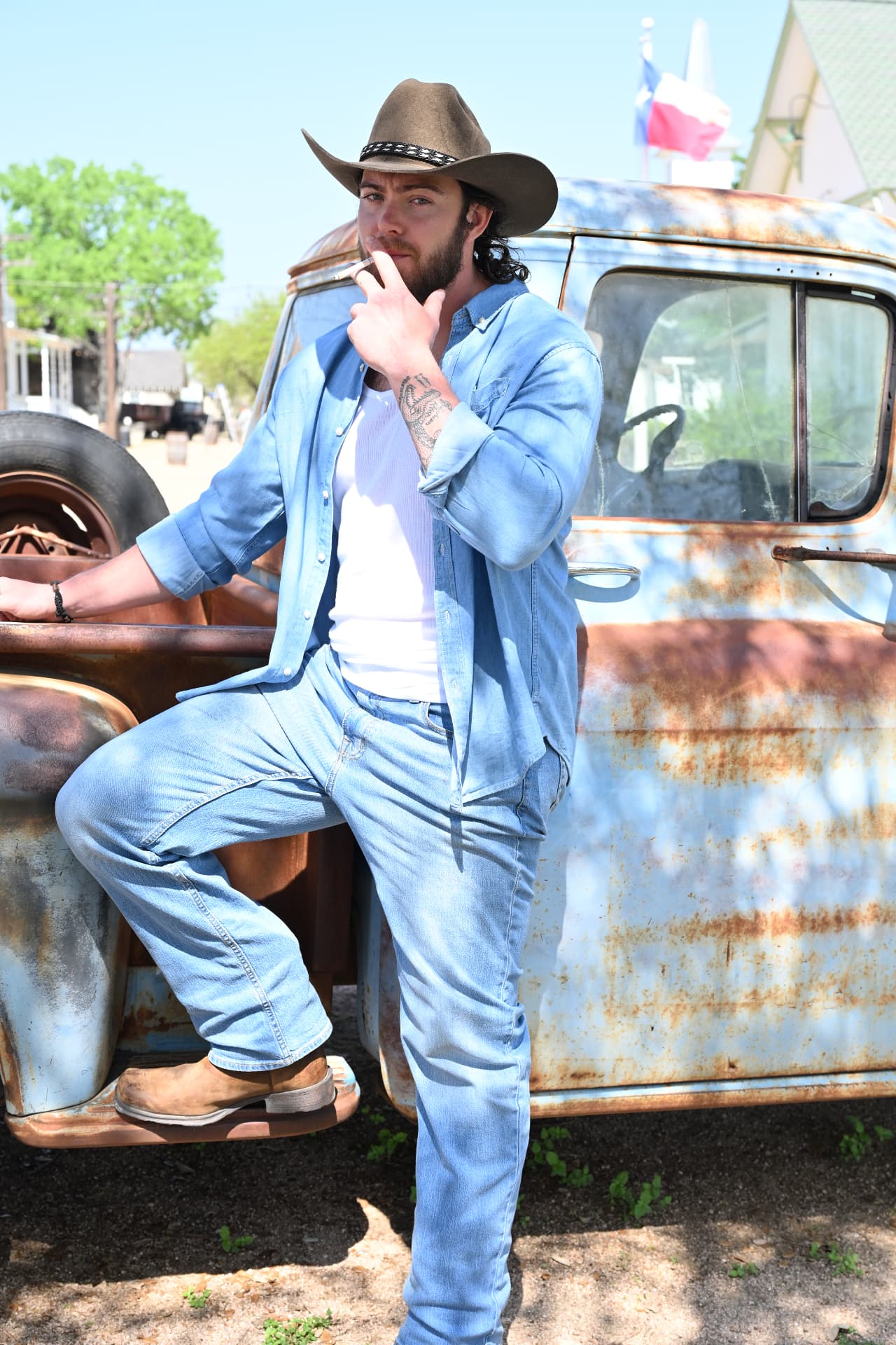 Jake Colletti full-length leaning against a rusted pickup truck in double denim and boots, Texas flag behind