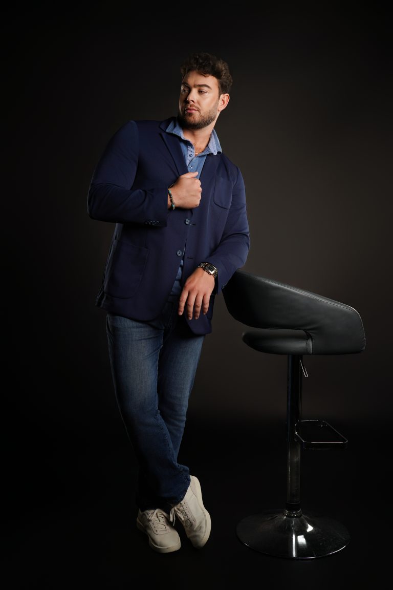 Jake Colletti standing full-length in a navy blazer, jeans and sneakers beside a bar stool, sophisticated dark studio look