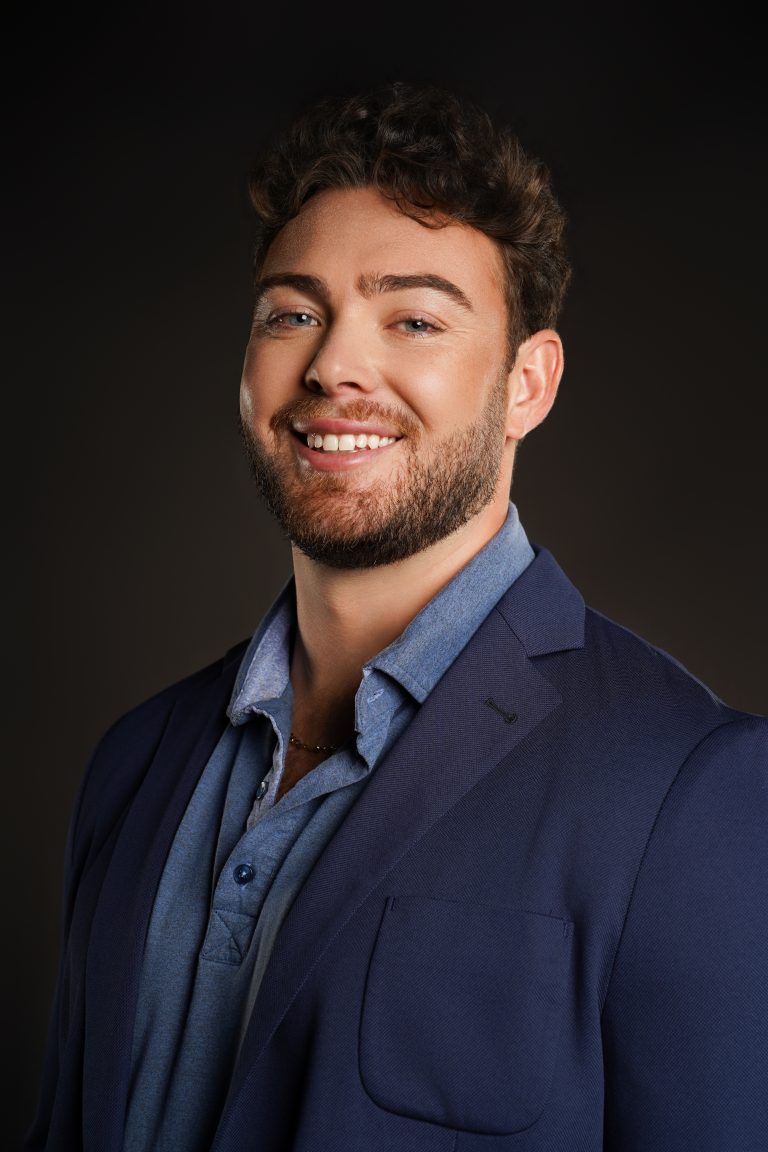 Jake Colletti theatrical headshot — smiling in a navy blazer over a chambray shirt against a dark studio backdrop