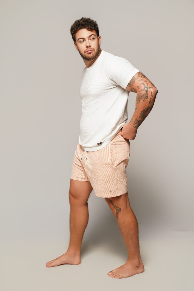 Jake Colletti full-length modeling shot in a white tee and striped swim shorts, barefoot on a grey studio backdrop, arm tattoos visible