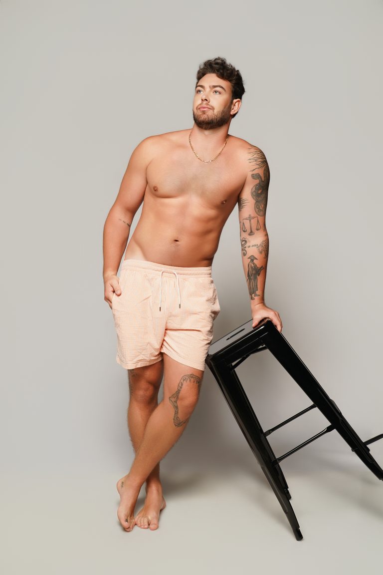Jake Colletti shirtless full-length physique shot in striped swim shorts leaning on a black stool, showing arm and leg tattoos