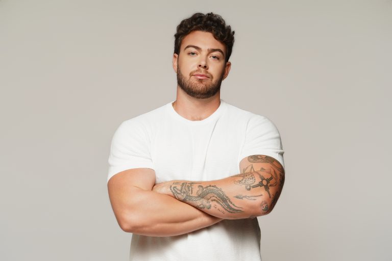 Jake Colletti commercial headshot in a white textured tee, arms crossed showing full left-arm tattoo coverage, grey studio backdrop