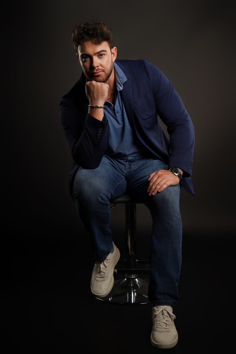 Jake Colletti seated on a stool in a navy blazer and jeans, hand to chin, dramatic dark studio lighting