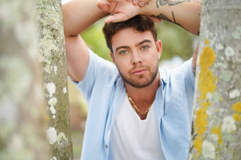 Jake Colletti outdoors in an open light blue shirt over a white tee, leaning between two lichen-covered tree trunks
