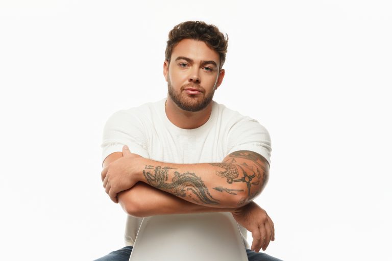 Jake Colletti seated in a white tee with arms crossed on a bright white studio backdrop, forearm tattoos visible