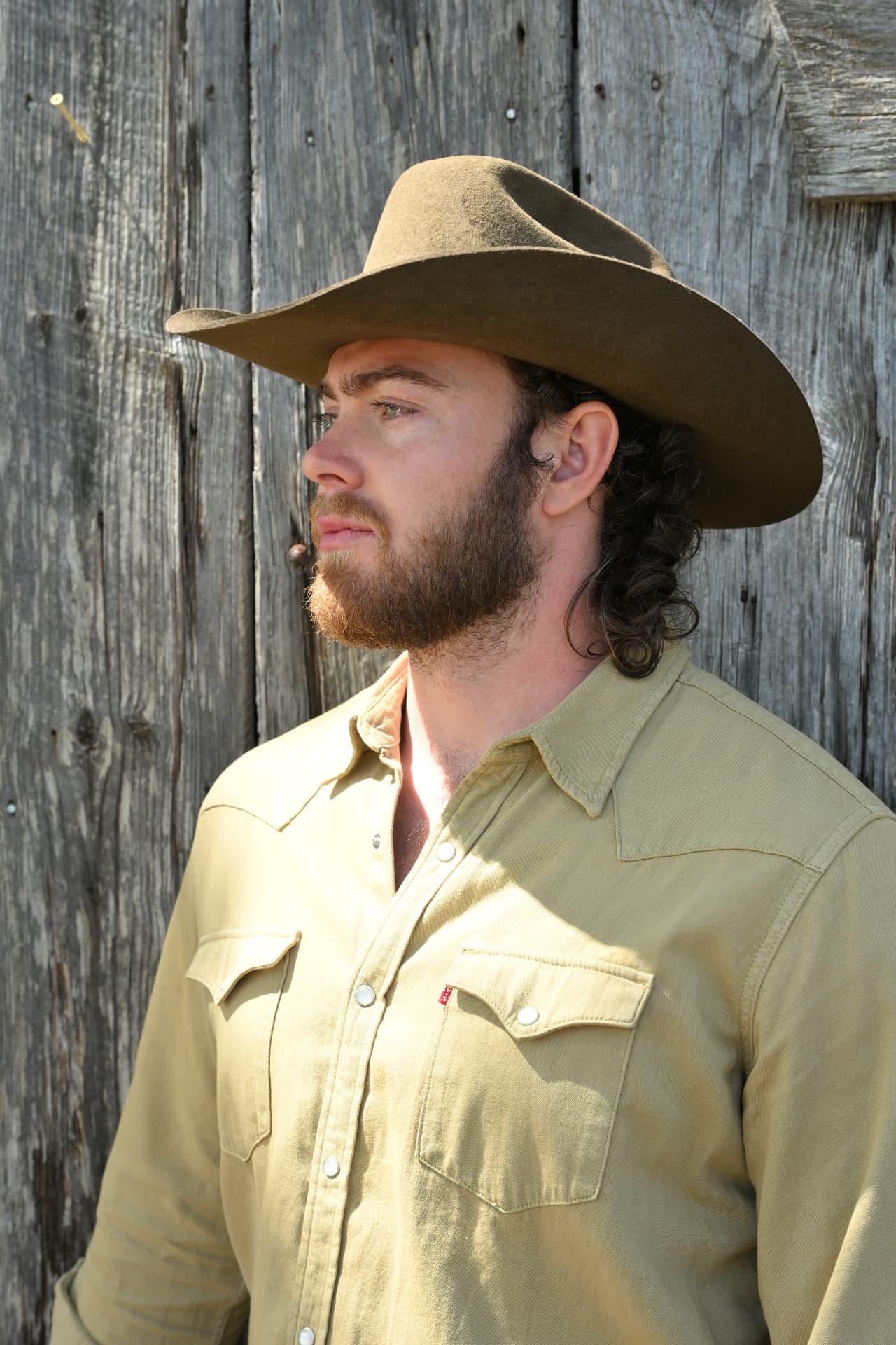 Jake Colletti rugged Western profile in a felt cowboy hat against a weathered barn wall, full beard and long hair