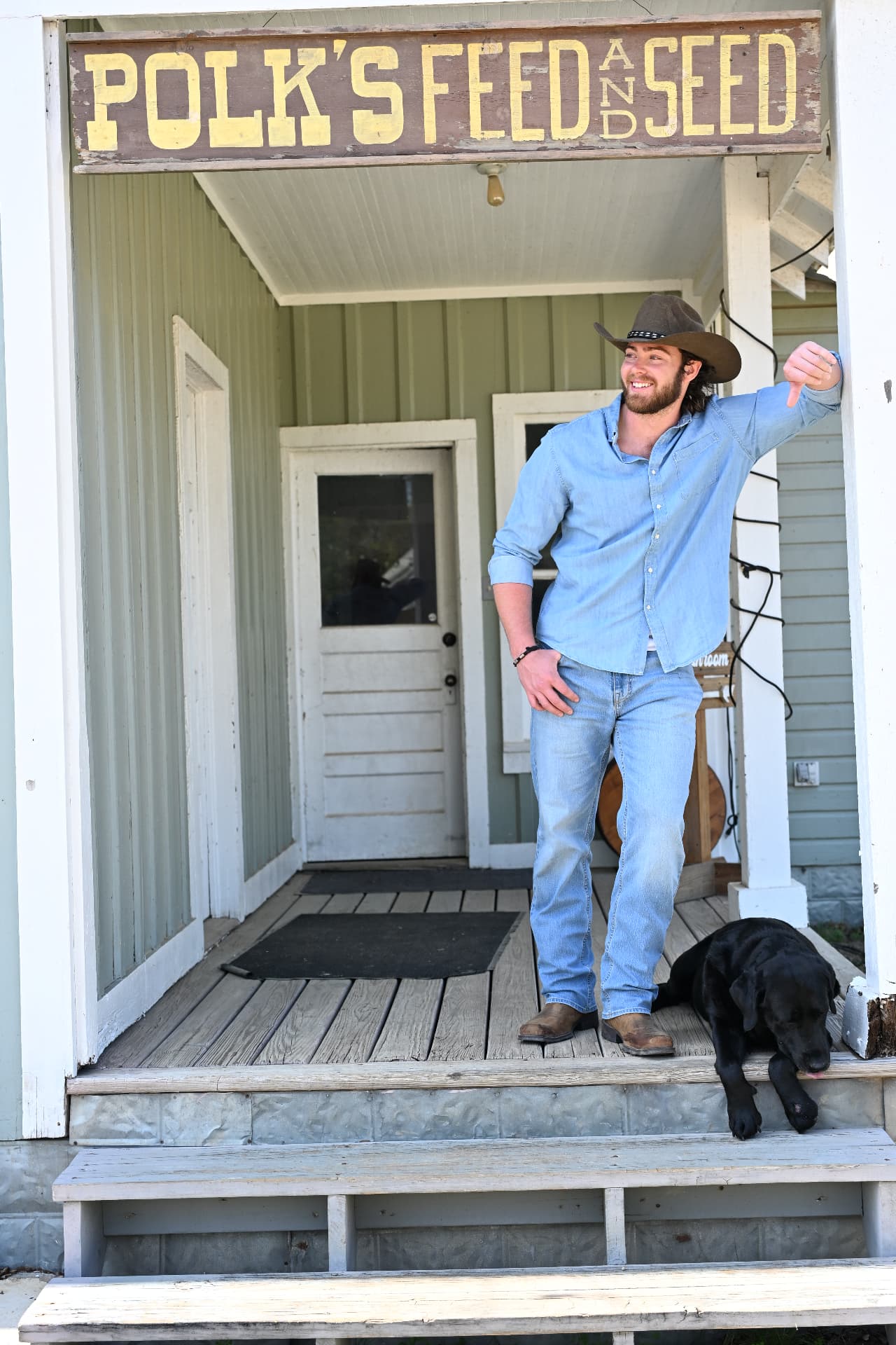 Jake Colletti smiling in a blue denim shirt and cowboy hat, leaning against a porch post with a black Labrador at his feet