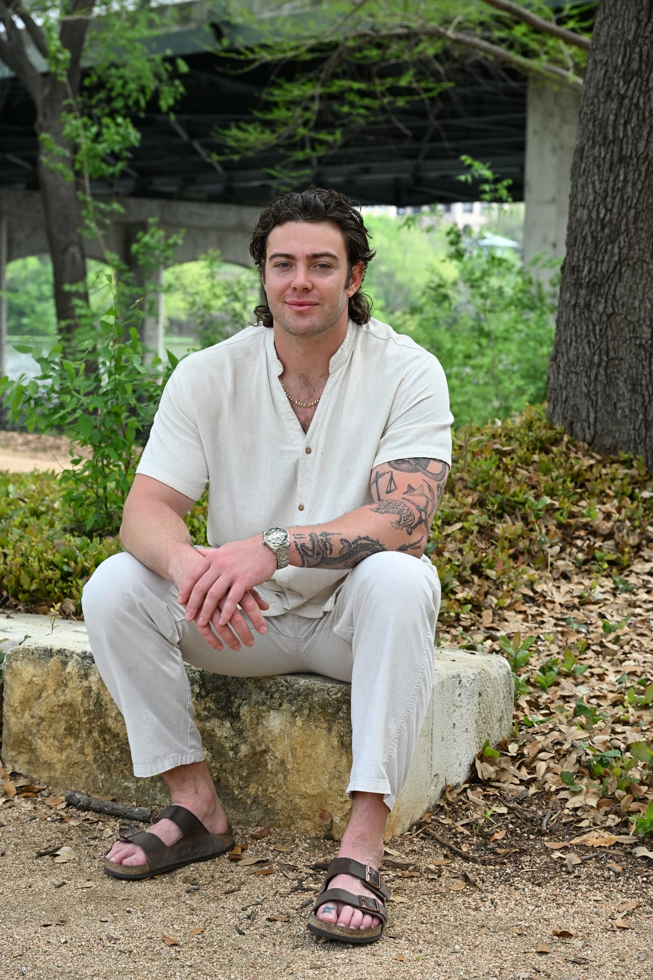 Jake Colletti editorial portrait in cream linen seated on stone under a bridge, showing arm tattoo coverage